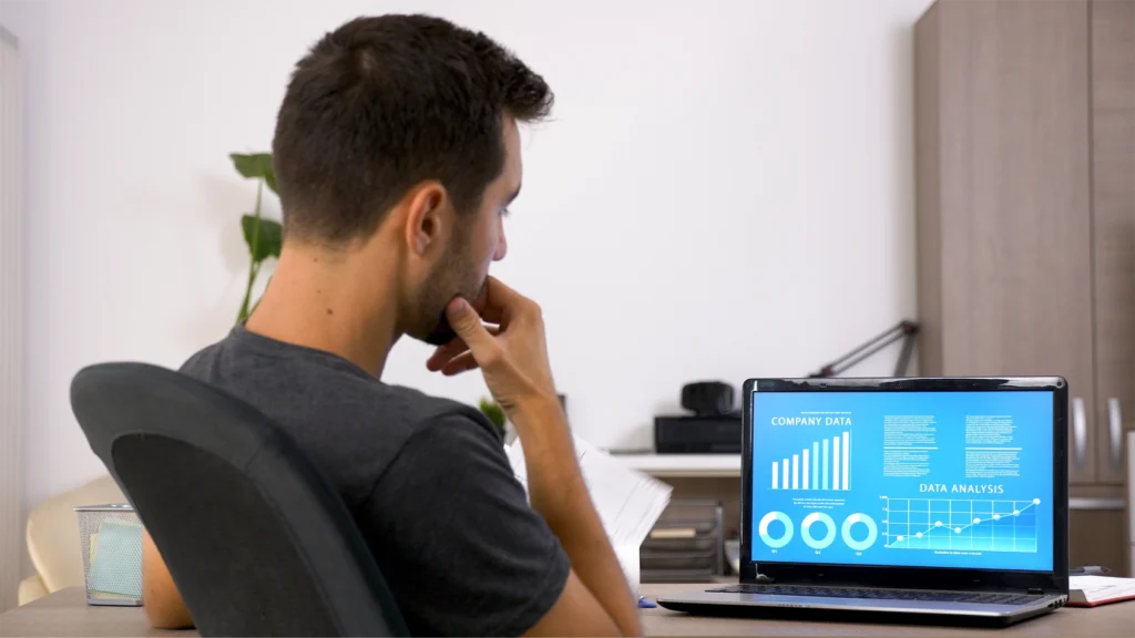 A young dark haired man sitting at a desk looking at a computer. On the screen is a data analytics dashboard.